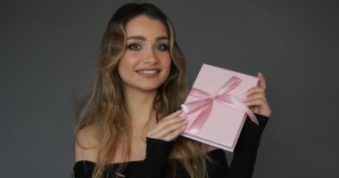 Charming young woman with flirtatious smile shows pink gift box with matching silk bow, hands it to camera. Studio grey background. Concept of beauty. Gift certificate. 4k footage.