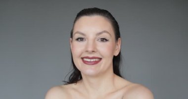 Close-up image of smiling woman with combed-back hair, who turns and tilts her head to demonstrate professional makeup. Concept of beauty. Studio grey background. Face and body care. 4k footage.