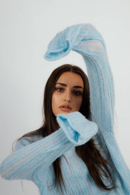 Kolları geniş, mavi kazaklı çekici bir kadın. Beyaz arkaplan, kadın stüdyo portresi. Kollarını kaldırmış, gündelik kıyafetlerle poz veren genç bir kadın. Moda, tarz, temel kıyafetler..