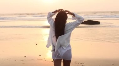 Beyaz tişörtlü, uzun dalgalı saçlı seksi Asyalı kadın ıslak kumda okyanusa ayak izleri bırakıyor. Dinlen ve rahatla. Güzellik ve sağlık. Turizm ve seyahat. Tam HD görüntüler.