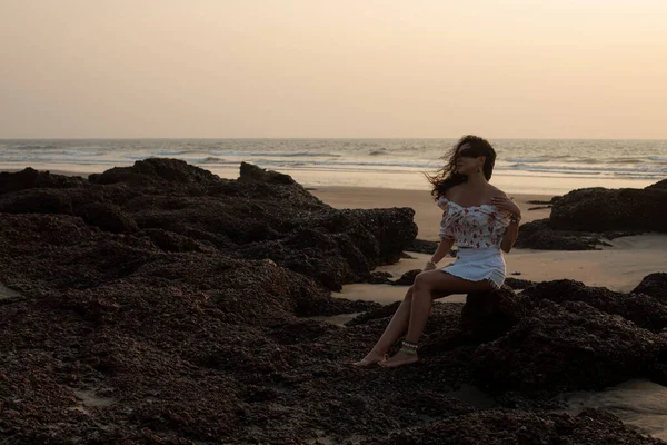 Young tanned woman with long hair fluttering in wind in short white skirt and summer blouse is sitting on ocean shore on rocks in sunset rays. Sunset on sea. Tourism and travel. Serene time.