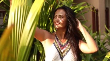 Saçında beyaz çiçekle gülümseyen yakışıklı Asyalı kadın lüks oteldeki tropikal bitki örtüsü arasında harika bir gün geçiriyor. Turizm ve seyahat. Egzotik cennet. Tam HD görüntüler.