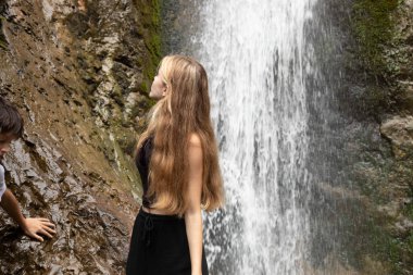 Genç, güzel, sarışın, uzun saçlı bir kadın dağlarda, küçük bir çocuğun kayalara tırmandığı doğal şelaleye doğru dönüyor. Kardeş seyahati, turist macerası ve keşfi.