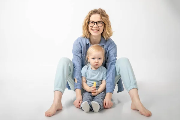 stock image Young charming red-haired woman sits on white background in studio and hugs her little daughter. They are dressed in clothes of same shades. Motherhood. Care and tenderness. Parents and children.