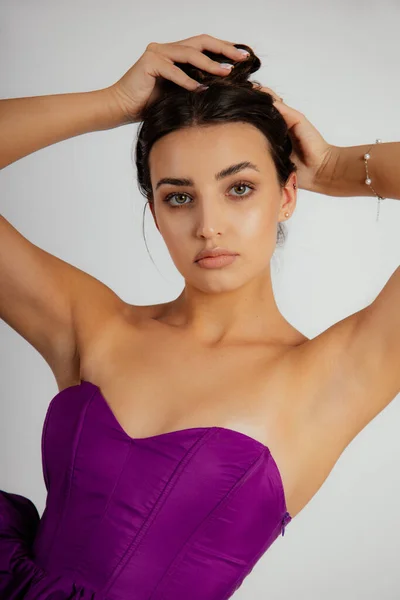 Vertical closeup young brunette woman in violet dress with bare shoulders, bracelet looking camera, posing with hands making hair bun on head in white studio. Smooth armpits, hair removing. Copy space