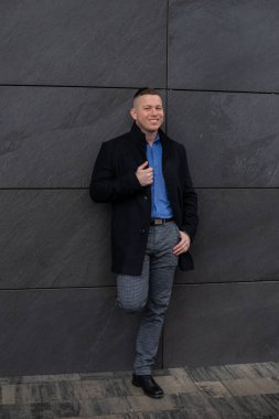 Siyah ceket giymiş, şehir binasının koyu gri duvarına karşı duran çekici genç bir mimarın tam boy görüntüsü. Kariyer ve başarı. Serbest çalışıyorum. Pozitif duygular.