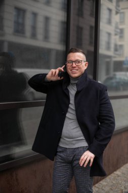 Şehir caddesinde bulutlu bir günde gri kazaklı ve siyah ceketli gülümseyen genç adamın portresi. İşletme ve girişimcilik. Serbest çalışıyorum. İletişim ve iletişim. 