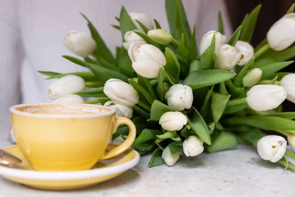 Yellow ceramic mug with cappuccino is on table in cafe, next to it lies luxurious bouquet of white tulips. Floral surprises. Romantic date. Spring mood. Positive emotions. Spring gifts.