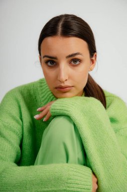 Uzun siyah saçlı, yeşil kazak giyen, pantolonu kalkmış bacaklarıyla oturan, dizlerini kucaklayan, çenesini elinde tutarak beyaz arka planda poz veren güzel bir kadının portresi. Dikey, stüdyo.