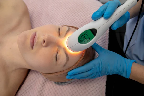 Relaxed female patient lying and getting rejuvenation laser facial procedures with laser nozzle with temperature data display in spa. Facial lifting contouring procedures for beauty. High angle view