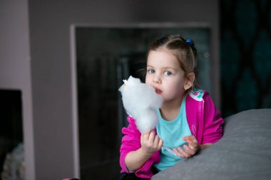 Meraklı, güzel, sarışın, neşeli, pamuk helva yiyen, çubukta pamuk helva yiyen ve kameraya bakan küçük kız. Çocuklar şekerlerle, şekerlerle eğlenir. Atıştırmalıklar ve sağlıksız şeker yiyecekler. Boşluğu kopyala