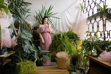 Ev serası organizasyonu. Omzundan pembe elbisesi olan çekici bayan çiçekçi serasında tropikal bitkilerin yanında duruyor. Hobi olarak. Çiçek işi. Bitkilerin satışı ve yetiştirilmesi. 