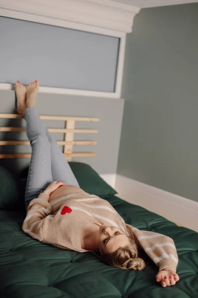 家の服を着た幸せな妊婦は 足を上げてベッドの上に横たわっています モダンなベッドルームのインテリアデザイン スタイリッシュな快適な家具 休憩所 若い母親 家庭のリラクゼーション — ストック写真