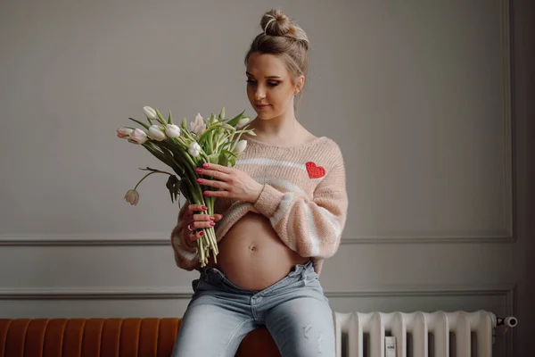 可愛い妊婦さんはリビングルームでソファに座っている新鮮な白いチューリップの花束を持っています インテリアデザイン 美しい春の花 ホーム女性写真 妊娠中 出産予定 若い母親 — ストック写真
