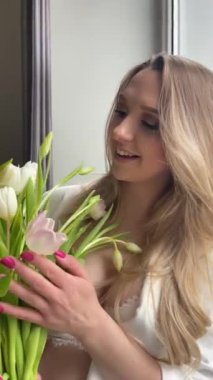 Taze kokulu lalelerle dolu mutlu hamile bir kadın. Pencere kenarında dikey kadın videosu. Beyaz iç çamaşırlı genç ve güzel bir bayan ve ev kıyafetleri çiçek aromalı. Tatil hediyesi.