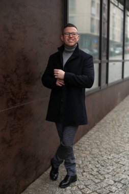 Gözlüklü, gri kazaklı ve siyah kaşmir ceketli başarılı gülümseyen adamın tam boy görüntüsü şehir caddesinde cam vitrinin yanında duruyor. İşletme ve girişimcilik. Kariyer ve başarı. 