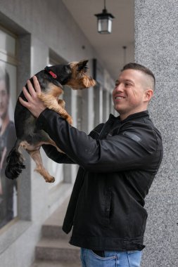 Kotlu ve siyah ceketli genç yakışıklı adam köpeğini kollarında tutuyor ve ona gülümsüyor. Evcil hayvanlar. Hayvanların bakımı ve bakımı. Köpeklerle yürümek. Pozitif duygular. 