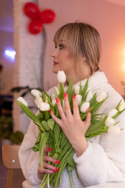 Young beautiful woman in white coat gently hugs large armful of white tulips, standing in festively decorated cafe. Floral surprises. Romantic date. Spring mood. Positive emotions.