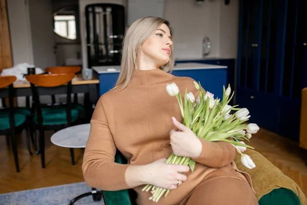 Pregnant young blonde with long hair in beige suit comfortably settled on sofa with bouquet of tulips at home. Modern business ladies. Self-sufficient women. Healthy pregnancy. Lifestyles.