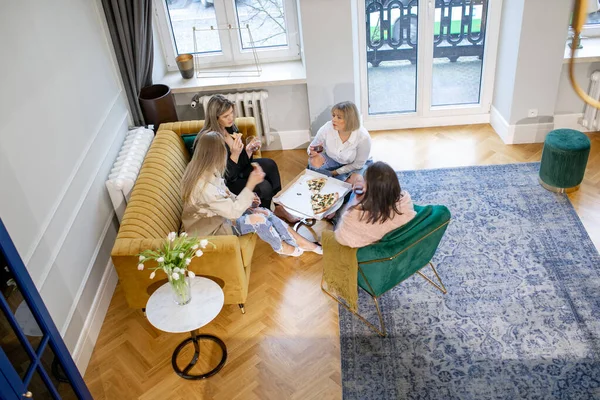 Group of women eat pizza from box, relax on sofa and talk in living room. Friends have dinner on home meeting. Female party, lunch break. Apartment interior design, from above view.