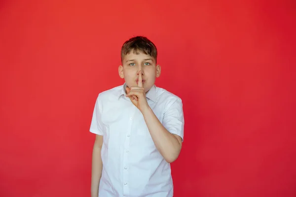Attention silence. Cute 12-year-old teenage boyt in white shirt with short sleeves presses his finger to his lips. Facial expressions and gestures. Red studio background. You cant make noise.