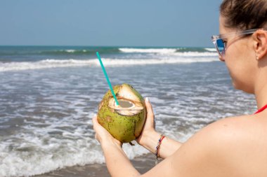 Tanınmayan genç bir kadın okyanusa bakıyor ve uzanmış ellerinde samanla hindistan cevizi soyuyor. Turizm ve seyahat. Yeni duygular. Mutluluk. Rahatlama. Anın tadını çıkarıyorum..