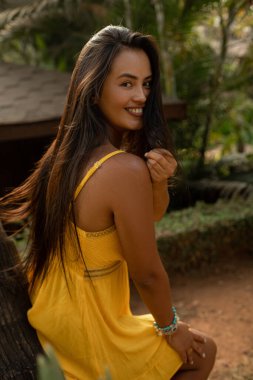 Kameraya gülümseyen parlak sarı yazlık elbiseli, uzun, gevşek saçlı çekici genç bir kadının portresi. Turizm ve seyahat. Tropik oteller. Dinlenmenin tadını çıkarıyorum. Bronzlaşma ürünleri. Cilt bakımı. 