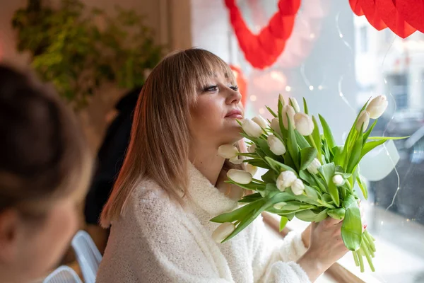 Beautiful young woman is sitting in cafe with luxurious bouquet of white tulips and dreamily looks out window on Valentines Day. Stylish women. Rest and relaxation. Live in present moment.