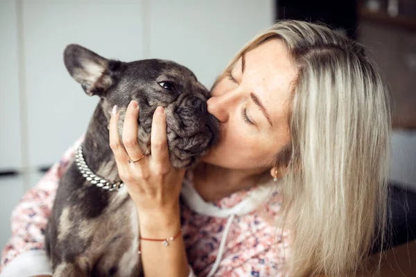 Love for your pets. Blonde woman he adores and kisses his French bulldog in cute muzzle. Maintenance and care of dog. Pets. Friendship and mutual understanding. Happy moments. Positive emotions.
