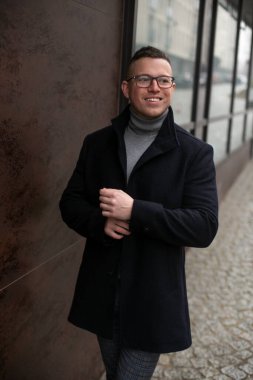 Gözlüklü, gri kazaklı ve siyah kaşmir ceketli başarılı gülümseyen adam şehir caddesinde cam vitrinin yanında duruyor. İşletme ve girişimcilik. Kariyer ve başarı. Bağımsız. 