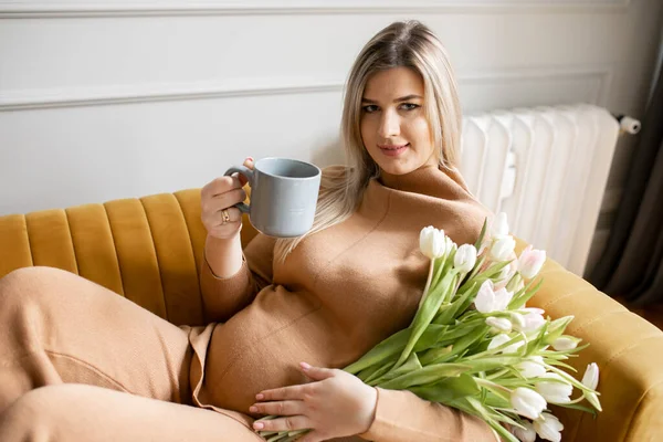 Smiling pregnant young blonde with long hair comfortably settled on sofa with bouquet of tulips and cup of tea at home. Modern business business ladies. Self-sufficient women. Healthy pregnancy.