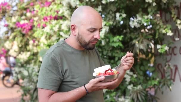 穿着灰色T恤的成熟秃头男人 吃着甜浆果 叉接着绿树吃着奶油 素食甜点 健康和维生素水果沙拉夏季 — 图库视频影像