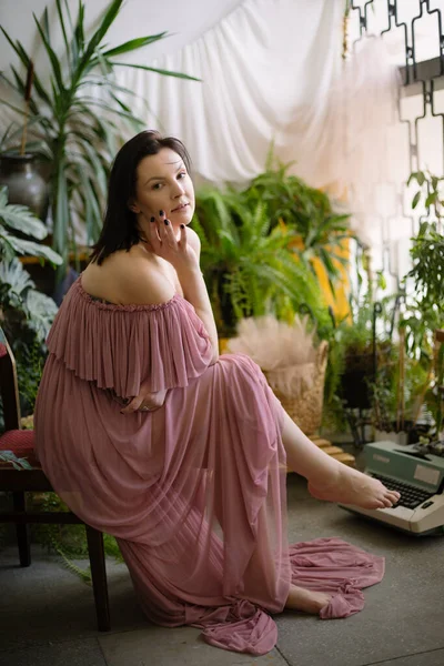 フラワービジネス ピンクのシフォンのドレスの魅力的な女性が冷静にカメラを見て 彼女の自宅の庭に裸足で座っている 趣味だ 温室だ 植物の栽培 植物用肥料 — ストック写真