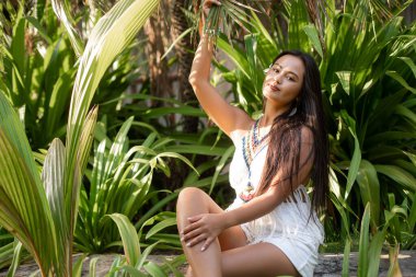 Büyüleyici esmer kadın poz veriyor, oturuyor ve sakin, yemyeşil bir parkta palmiye ağaçlarında dinleniyor. Doğayla çevrili huzurlu ve rahatlatıcı bir tatil arıyorum. Yaz tatili. Boşluğu kopyala