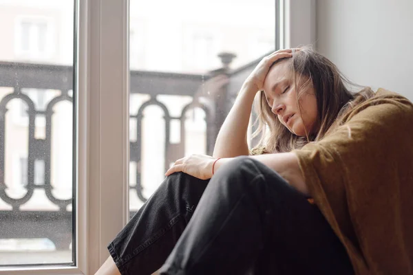 stock image Portrait of middle-aged desperate woman with long dark hair sitting with closed eyes on floor near window, putting hand on forehead, suffering from problems. Stress, depression, negative emotions.