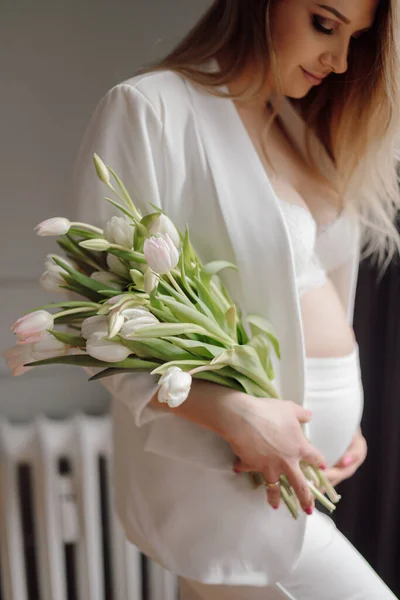 面带微笑的孕妇身穿白色紧身衣 站在窗前 捧着一束白色郁金香花 望着肚子的画像 — 图库照片