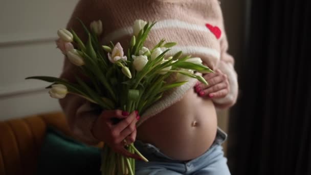 ジーンズの適用とベージュのセーターを着て認識できない若い妊婦は 裸の腹を撫で触れ 白いチューリップの花束を保持する 母親と妊娠期待愛 — ストック動画