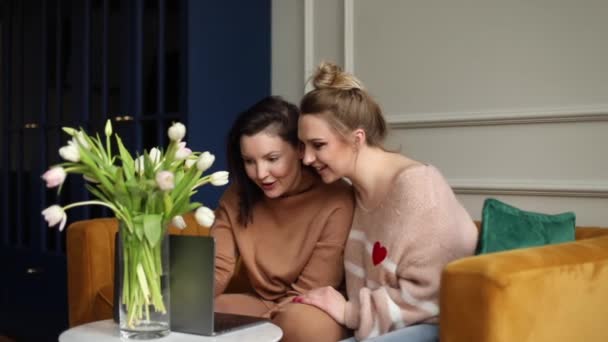 两位快乐的年轻女同事坐在花瓶里白色郁金香花束旁的黄色沙发上 看着灰色笔记本电脑的屏幕 笑着上网购物 交流工作 — 图库视频影像