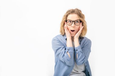 Şok olmuş, kısa dalgalı saçlı, ağzı açık, gözlük takan, kot gömlek giyen, elleri beyaz arka planda olan mavi pantolonlu genç bir kadının portresi. Aşırı duygusal, hisler..