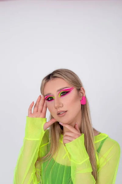 Face of female model with bright neon makeup. Beautiful blonde with long straight hair and fashionable makeup. Artistic designer makeup. Model in studio on light background. Beauty and freshness.