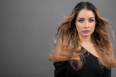Kadın saçı çıkıyor. Güzel, genç, dalgalı saçlı, omuzları açık bir manken stüdyoda gri arka planda kameraya bakıyor. Profesyonel makyöz. Kadın saçı çıkıyor.. 