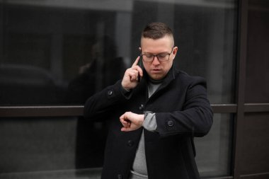 Siyah paltolu ve gözlüklü yakışıklı adam telefon ediyor. Bina duvarının yanındaki bilek saatine bakıyor. Meşgul bir iş adamının portresi. Zarif orta yaşlı insan. Sıradan erkek kıyafetleri. Çalışma çağrısı.