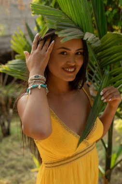 Palmiye yaprağının altındaki sarı elbiseli güzel esmer kadının portresini kapat, kameraya bak ve yeşil orman düzenindeki saçlara dokun. Tropik yaz seyahati ve tatildeki macera