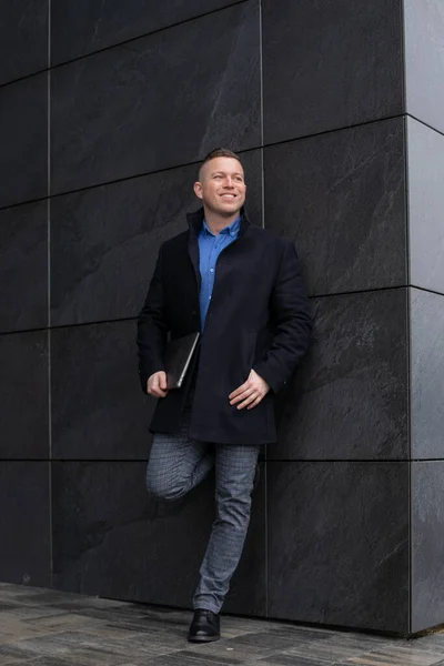 Handsome smiling man in black coat standing near building wall with laptop. Portrait of happy working business man. Elegant middle aged person. Casual male outwear. Working lifestyle.