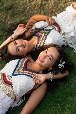 Üst Manzara. Saçlarında beyaz çiçekler olan iki çekici Asyalı kadın zarif bluzlar giyip lüks otellerde çimlere uzanıyor ve gülümsüyor, kameraya bakıyor. Turizm ve seyahat. Dinlen ve eğlen.. 