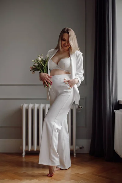 Smiling blonde with loose long hair in elegant white maternity suit stands barefoot on parquet floor in expensive interior in gray tones. Maternity insurance and protection.