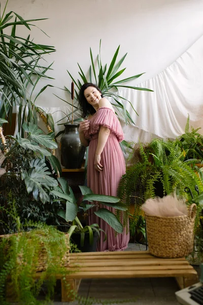 Charming landscape designer woman in pink off-the-shoulder dress enjoys beautiful plants in her greenhouse, energizes herself. Hobby. Flower business. Plant care. Sale and cultivation of plants.