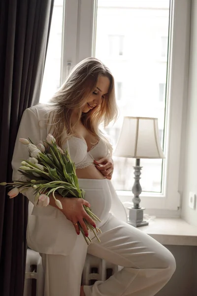 チューリップの花束と白いスーツの妊娠中の若い女性は窓の近くに立って 穏やかな笑顔で彼女の胃に触れます 誕生を待っている 育児と母親 愛と優しさ — ストック写真