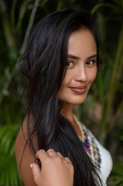 Dikey olarak, uzun saçlı esmer kadın kameraya gülümsüyor ve yaz tatilinde parkta yeşil tropikal bitkilere dokunuyor. Tropik cennete tek başına yolculuk planlamak gevşemek, keşfetmek