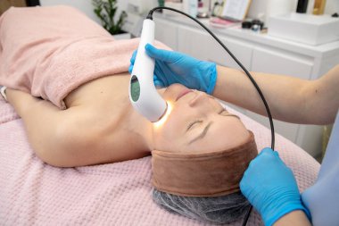 Rahatlamış bir kadın klinikte yatarken lazer yüz tedavisi görüyor. Genişleme için hafif radyasyon teknolojisi. Yüz gerdirme, kırışıklık önleyici ultrason ve radyofrekans teknolojisi.